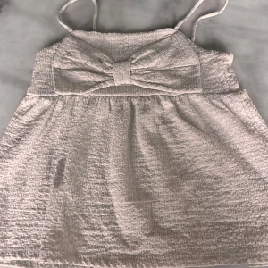 Vit linnetopp med rosett från Kappahl - Supersöt vit linnetopp med smala axelband och en stor rosett framtill. Toppen är tillverkad i ett mjukt material som känns luftigt. Perfekt för varma dagar och ger en gullig och trendig look. 💗