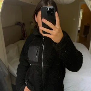 Svart teddyjacka med dragkedja - Mysig svart teddyjacka med hög krage och hel dragkedja framtill. Jackan har en praktisk bröstficka med dragkedja i kontrasterande material och två sidofickor. Perfekt för kyliga dagar och riktigt skön att bära.