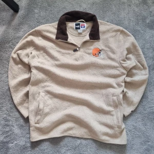 Beige fleece-tröja Puma clevland browns - Säljer en beige fleece-tröja från Puma NFL med broderad logga och Cleveland Browns hjälm på bröstet. Tröjan har mörkbrun krage, half zip och två sidofickor. Materialet är mjuk polyester och modellen är långärmad, perfekt för kyliga dagar.