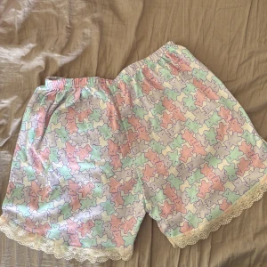 Pastelliga shorts med nallemönster och spets - Supergulliga shorts med pastellfärgade nallar i rosa, mintgrönt, lila och ljusgult. De har elastisk midja och söt spetskant längst ner. Perfekta för mysiga dagar hemma eller som pyjamasshorts. Mjukt bomullsmaterial som känns skönt mot huden.