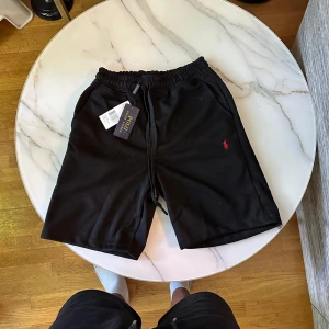 Svarta shorts från Polo Ralph Lauren - Svarta shorts från Polo Ralph Lauren med elastisk midja och snörning. Klassisk röd broderad logga på ena benet. Sköna och stilrena, perfekta för sommaren. Tillverkade i mjuk bomull för en chill vibe.