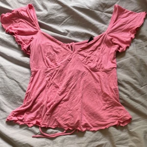 Rosa knyttopp från Gina Tricot - Supersöt rosa topp från Gina Tricot med omlottdesign och knytband där bak. Toppen har korta volangärmar och vågig kant nertill. Mjuk och lätt i bomull, perfekt för en somrig look. Säljer då den inte längre kommer till användning!