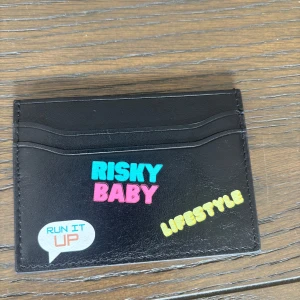 Risky Lifestyle svart korthållare - Svart korthållare i skinn från Risky Lifestyle med färgglada tryck som 'RISKY BABY', 'LIFESTYLE' och 'RUN IT UP'. Slimmad design med flera kortfack och präglad R-logga på baksidan. Perfekt för dig som vill ha en unik och stilren accessoar.