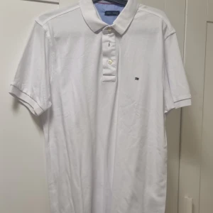 Vit pikétröja Tommy Hilfiger XL - Klassisk vit pikétröja från Tommy Hilfiger i storlek XL. Tröjan har korta ärmar, krage och knäppning med tre knappar framtill. Liten broderad logga på bröstet. Perfekt för en clean och stilren look.