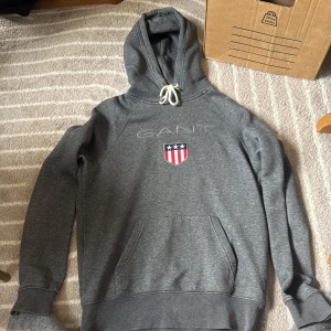 Grå hoodie från GANT med brodyr - Snygg grå hoodie från GANT med broderad logga och sköld på bröstet. Tröjan har huva med vit snörning och en stor magficka. Nypris: 1200