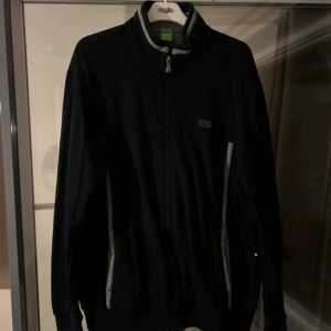Svart trackjacka från Hugo Boss - Svart trackjacka från Hugo Boss med gråa detaljer längs sidorna och på kragen. Jackan har dragkedja hela vägen, ribbade muddar och en diskret Boss-logga på bröstet. Perfekt för en sportig och clean stil.