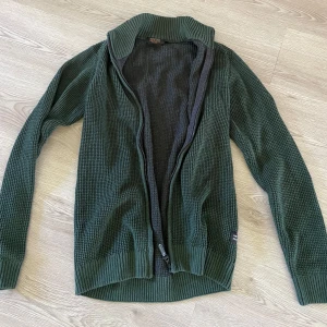 Mörkgrön stickad kofta från Indicode - Säljer en mörkgrön stickad kofta från Indicode i storlek M. Koftan har grovstickad struktur, hög ribbad krage och hel dragkedja framtill. Perfekt för lager-på-lager och chillare dagar. Snygg och enkel att matcha med jeans eller chinos.