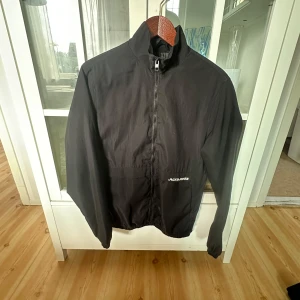 Svart vindjacka från Jack & Jones S - Svart vindjacka från Jack & Jones i storlek S. Jackan har hög krage, hel dragkedja framtill och diskret logga på bröstet. Tillverkad i lätt polyester med elastisk nederkant och ärmslut. Perfekt för dig som gillar en clean och enkel stil.