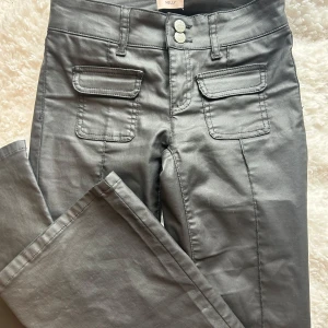 Svarta bootcut-byxor från Nelly - Snygga svarta bootcut-byxor från Nelly med två framfickor med lock och dubbla knappar i midjan. Byxorna har en lätt glansig finish och markerade sömmar framtill. Perfekta för dig som vill ha en trendig och stilren look.