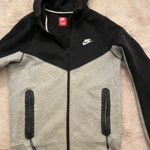 Nike hoodiejacka svart/grå S - Nike hoodiejacka i svart och ljusgrått med dragkedja hela vägen, två fickor med dragkedja och klassisk Nike-logga på bröstet. Jackan har huva och är tillverkad i mjuk bomullsblandning, perfekt för chill eller träning.