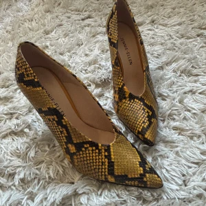 Gula pumps med ormmönster från Jennie-Ellen - Snygga pumps från Jennie-Ellen i gul och svart ormmönstrad skinnimitation. Skorna har spetsig tå och hög klack, vilket ger en edgy och trendig look. 