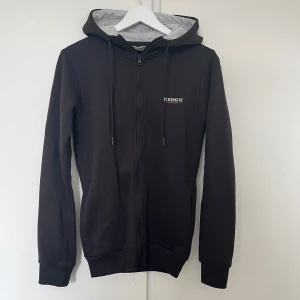 Svart hoodie från McKenzie XS - Svart hoodie från McKenzie i storlek XS. Tröjan har dragkedja framtill, snörning i huvan och grått innerfoder i luvan. Den tajt. Liten vit logga på bröstet. Perfekt för chill dagar eller när du vill ha en enkel streetstyle.