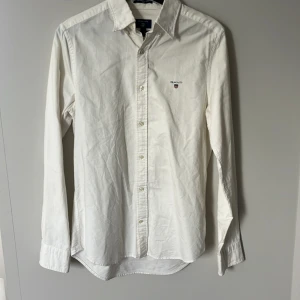 Vit skjorta från GANT - Vit Oxfordskjorta från GANT i slim fit-modell, storlek S (37/38). Klassisk design med knappar framtill, lång ärm och broderad logga på bröstet. 