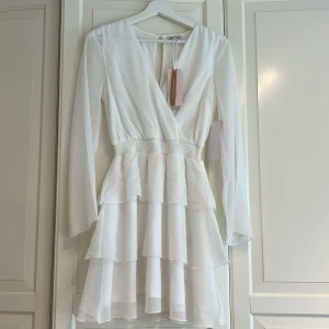 Vit omlottklänning med volanger - Supersöt vit omlottklänning med v-ringning, långa ärmar och flera lager volanger i kjolen. Smockad midja ger en snygg siluett och det tunna, luftiga materialet gör den perfekt för varmare dagar. I storlek 36!