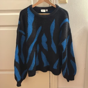 Svart-blå stickad tröja från VILA - Cool stickad tröja från VILA i svart med blått zebramönster. Oversized passform med långa ärmar och rund hals. Mjuk och fluffig känsla, perfekt för att ge din outfit en edgy vibe. Snygg att styla med jeans eller kjol.