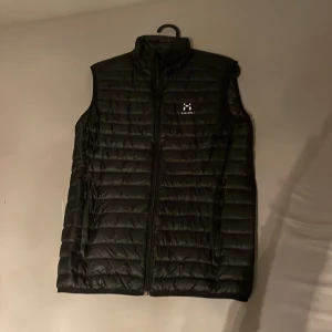 Svart dunväst från Haglöfs - Säljer en svart dunväst från Haglöfs med quiltad design och hög krage. Västen har dragkedja framtill och diskret logga på bröstet. Perfekt att slänga över en hoodie för extra värme. Klassisk och stilren look som funkar till det mesta.
