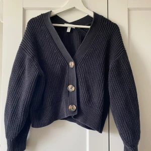 Svart stickad kofta från H&M - Mysig svart stickad kofta från H&M i storlek XS. Koftan har grov ribbstickning, djup v-ringning och stora träknappar framtill. Långärmad och lite boxig passform som ger en chill vibe. Perfekt att slänga över en t-shirt eller linne.