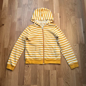 Gulvit randig hoodie från H&M - Säljer en fräsch hoodie från H&M i storlek 134/140. Tröjan är gul och vit med snygga horisontella ränder, har huva och dragkedja framtill samt två fickor. Muddar vid ärmslut och nederkant ger en chill vibe. Perfekt för dig som gillar färg och mönster!