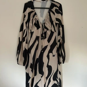 Zebramönstrad långklänning från NA-KD - Långklänning från NA-KD med zebramönster i svart och beige. Klänningen har djup v-ringning med justerbar snörning framtill och långa puffärmar med resår vid ärmslut. Materialet är lätt och skönt. Använd 1 gång.