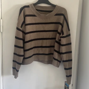 Beige och svart randig stickad tröja - Säljer en beige stickad tröja med svarta horisontella ränder. Tröjan har en rund halsringning, ribbade muddar och är långärmad. Den är loose fit och har en chill, oversized vibe som passar perfekt till jeans eller leggings. Den är lite kortare i modellen. Vet inte vart den är ifrån tyvärr. Nypris 129. Lite nopprig.
