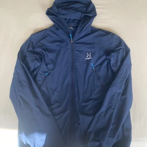 Blå vindjacka från Haglöfs med huva - Säljer en blå vindjacka från Haglöfs i storlek L men sitter mer som S eller M. Jackan har huva, två snedställda fickor med dragkedja och hel dragkedja framtill. Perfekt för uteliv och äventyr, lätt och smidig att bära med sig. Klassisk Haglöfs-logga på bröstet. 