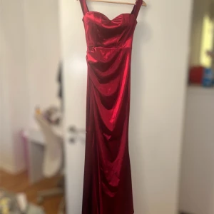 Röd Vintage långklänning med breda axelband - Snygg röd Vintage långklänning med breda axelband och figurnära passform. Klänningen har ett glansigt satinliknande material och ett hjärtformat bröstparti som ger en elegant look. Perfekt för bal eller fest, och den har en stilren design utan extra detaljer.