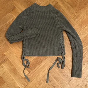 Grå ribbad stickad tröja med snörning - Säljer en grå stickad tröja med ribbad struktur och snygg snörning i sidorna. Tröjan har lång ärm och en högre krage som ger en cool vibe. Perfekt för dig som gillar detaljer och vill ha något som sticker ut lite extra.