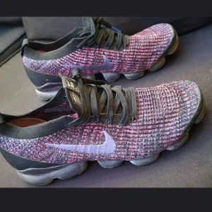 Nike Vapormax Flyknit sneakers - Nike Vapormax Flyknit sneakers med stickad ovandel i svart, rosa och blå toner. Snyggt mönstrad mesh med svart snörning och tydlig Nike-logga på sidan. Sulan är platt och har den klassiska Vapormax-bubblan för extra komfort. Perfekt för dig som gillar sportig och modern stil. Se bilder för storlek och skick. 