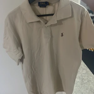 Beige Polo t-shirt från Ralph Lauren - Snygg beige t-shirt från Polo Ralph Lauren med klassisk krage och korta ärmar. Ikoniska broderade loggan på bröstet. Perfekt för dig som gillar stilrena och tidlösa plagg. Mjuk bomull som känns skön mot huden.