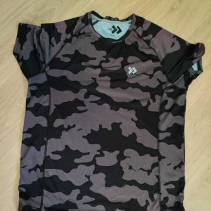 Svart och grå camo t-shirt - Snygg t-shirt från Daiwa med svart och grått camouflage-mönster. Kortärmad och tillverkad i lätt polyester som andas, perfekt för träning eller chill. Logga på bröstet ger en sportig vibe. Passar dig som gillar streetstyle och funktion.