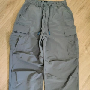 Grå cargopants med snörning - Snygga grå cargopants med bred passform och praktiska sidofickor. Byxorna har elastisk midja med snörning och är perfekta för en avslappnad streetstil. Tillverkade i ett mjukt bomullsmaterial som känns skönt mot huden.