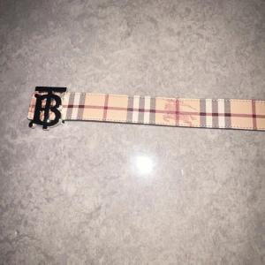 Burberry bälte med  Omvänd - Säljer två snygga Burberry-bälten med klassiskt rutigt mönster och TB-logga i metall. Ett bälte är beige med röda och svarta rutor, det andra är helsvart med diskret rutmönster. Båda har präglad Burberry-ryttare och är tillverkade i skinn.