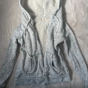 Vintage Hollister zip up - Mysig ljusgrå teddyjacka från Hollister med broderad logga på bröstet. Jackan har huva med snörning, dragkedja framtill och två fickor. Insidan är fodrad med fluffig teddy för extra värme. Perfekt för kyliga dagar.