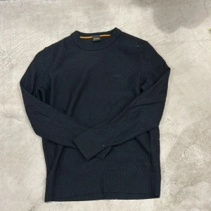 Svart stickad tröja från BOSS - Svart stickad tröja från BOSS med diskret broderad logga på bröstet. Rund hals med orange detalj på insidan av kragen. Tröjan har långa ärmar och ribbade muddar vid ärmslut och nederkant. Perfekt för dig som gillar stilrena och klassiska plagg.