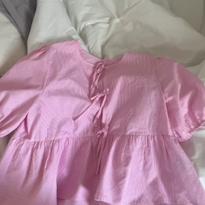 Rutig rosa blus med knyt bak - Supersöt rosa blus med små vita rutor och tre knytband där fram. Puffiga korta ärmar och volang nedtill. Klippt av lapparna så kommer inte ihåg vart den kommer ifrån. Storleken är iallafall i S. Använd endast en gång!💓