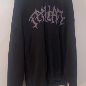 Svart hoodie med lila tryck - Svart hoodie från Weekday med ett fett lila tryck på bröstet i coolt grafiskt typsnitt. Tröjan har huva, lång ärm och en klassisk känguruficka framtill. Perfekt för dig som gillar streetwear och vill sticka ut med en unik design.