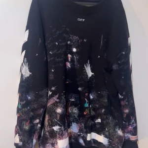 Off-White svart sweatshirt med tryck - Svart sweatshirt från Off-White med ikoniska vita pilar och ränder på ärmarna. Tröjan har färgstänk och konstnärligt mönster över hela plagget, samt ribbade muddar. Perfekt för dig som gillar streetwear och vill sticka ut med en unik design.