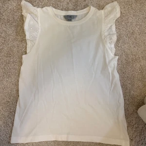 Vit ärmlös topp med volang  - Supersöt vit topp från Ullared med broderade volangärmar i spets. Toppen är ärmlös och har en rund halsringning. Storlek 134/140💓