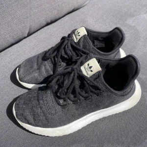 Svarta Adidas sneakers med vit sula - Svarta Adidas sneakers med klassisk vit sula. Skorna har snörning och en mjuk, flexibel känsla. Adidas-logga på plösen i beige och svart. Perfekta för dig som gillar en clean och sportig look. Använda några gånger men är i bra skick, vg se bilder för slitage. OBSERVERA att storleken är 36 3/4 i storlek. Jag har vanligtvis 37 i storlek och de passar bra.