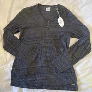 Mörkgrå långärmad tröja från edc - Snygg mörkgrå långärmad tröja från edc med diskreta ränder och tre knappar vid v-ringningen. Mjuk och skön bomullskänsla, perfekt för chill dagar. Passar dig som gillar enkel och stilren look.