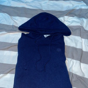 Mörkblå stickad hoodie från Bvaldi - Mörkblå stickad hoodie från Bvaldi med broderad logga på bröstet. Tröjan har huva med snörning och är gjord i ett mjukt stickat material som känns riktigt skönt. Perfekt för dig som gillar en chill och stilren look. Passar runt 165-175