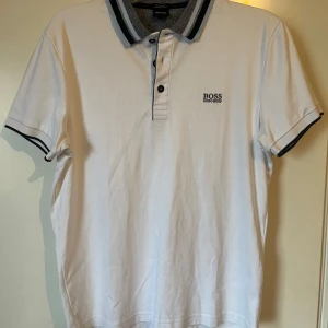 Vit Hugo Boss pikétröja M - Vit pikétröja från Hugo Boss i lyxig bomull. Tröjan har korta ärmar, klassisk krage med grå och svarta ränder, samt knappar framtill. Diskret Boss-logga på bröstet och snygga kontrastdetaljer vid kragen och ärmsluten. Perfekt för en clean och stilren look. Använd en gång ser ut som ny