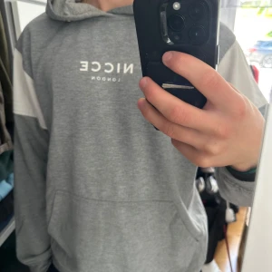 Grå hoodie från Nicce London - Säljer en grå hoodie från Nicce London med vita detaljer på ärmarna och tryckt logga på bröstet. Tröjan har huva, känguruficka och långa ärmar. Perfekt för chill dagar och streetstyle. Jag är 181cm och 75kg
