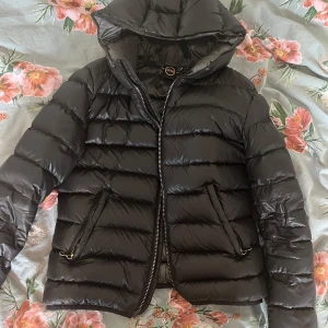 Colmar jacka  - Snygg svart dunjacka med huva och dragkedja framtill. Jackan har en quiltad design och långa ärmar, perfekt för kyligare dagar. Den har en normal passform och är både stilren och funktionell. 