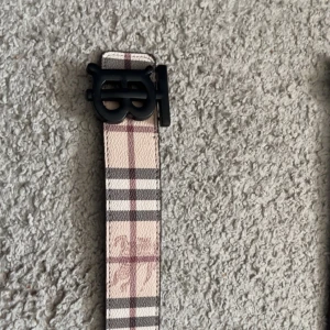 Burberry bälte - Snyggt bälte från Burberry i klassiskt rutigt mönster med beige, svart och vinröda toner. Bältet har en svart matt spänne med Burberrys logga och är gjort i skinn. Perfekt accessoar för att lyfta din outfit med en lyxig touch.