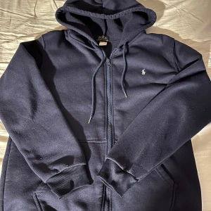 Mörkblå hoodie från Polo Ralph Lauren - Snygg mörkblå hoodie från Polo Ralph Lauren med dragkedja och huva. Tröjan har långa ärmar, ribbade muddar och klassisk Polo-logga broderad på bröstet. Perfekt för chill dagar och enkel att matcha med jeans eller joggers.