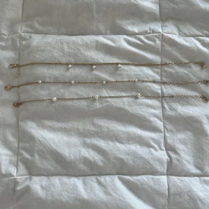 Guldiga armband med pärldetaljer - Tre tunna ankletbracelets i guldton med små vita pärlor. Två av armbanden har en tunn kedja med jämnt placerade pärlor, medan det tredje har en lite grövre kedja och tre pärlor som hänger. Perfekta för att mixa och matcha för en trendig look.