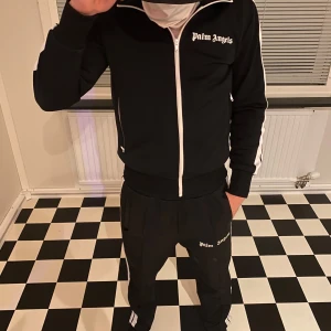 Palm Angels svart track jacket - Svart track jacket från Palm Angels med vita ränder längs ärmarna och vit dragkedja. Jackan har hög krage, ribbade muddar och logga på bröstet. Perfekt för en sportig och trendig look.