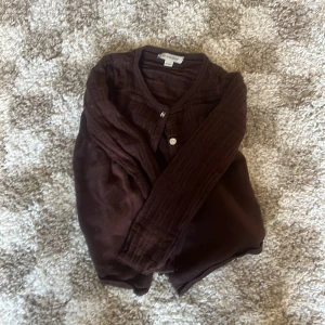 Brun kofta från Burberry, 8Y/128cm - Mörkbrun kofta från Burberry i stickat material med diskret rutigt mönster. Koftan har långa ärmar, knappar framtill och klassisk design. Perfekt för dig som gillar stilrena och tidlösa plagg med en lyxig touch.