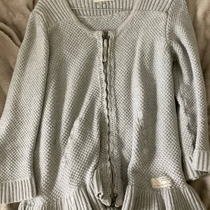 Ljusgrå kofta från Odd Molly - Superfin ljusgrå kofta från Odd Molly med stickad struktur, dekorativa virkade detaljer längs knäppningen och bred ribbad kant nedtill och vid ärmslut. Koftan har lång ärm och en klassisk passform som är både snygg och bekväm.